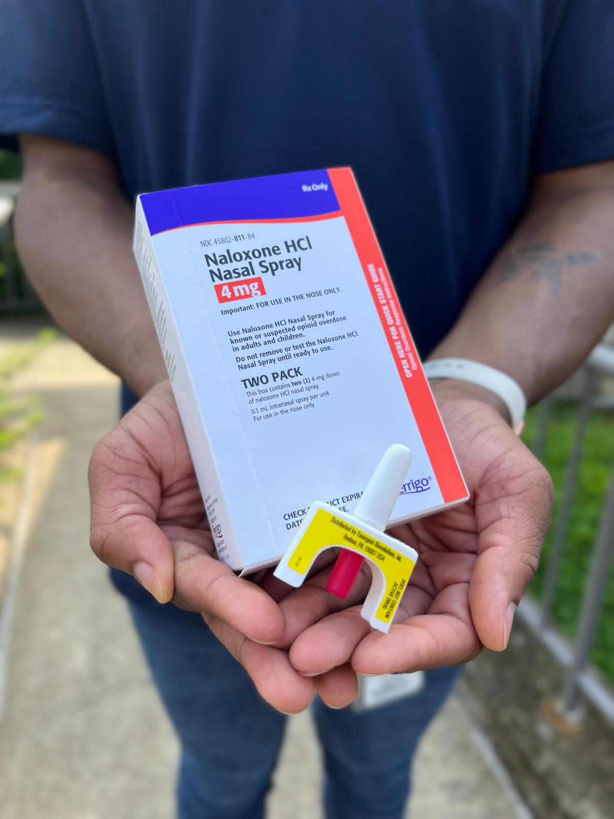 Close-up shot of two hands cupped together in front of a man's torso holding a box of Naloxone Nasal Spray and the nasal spray device used for Naloxone administration. 