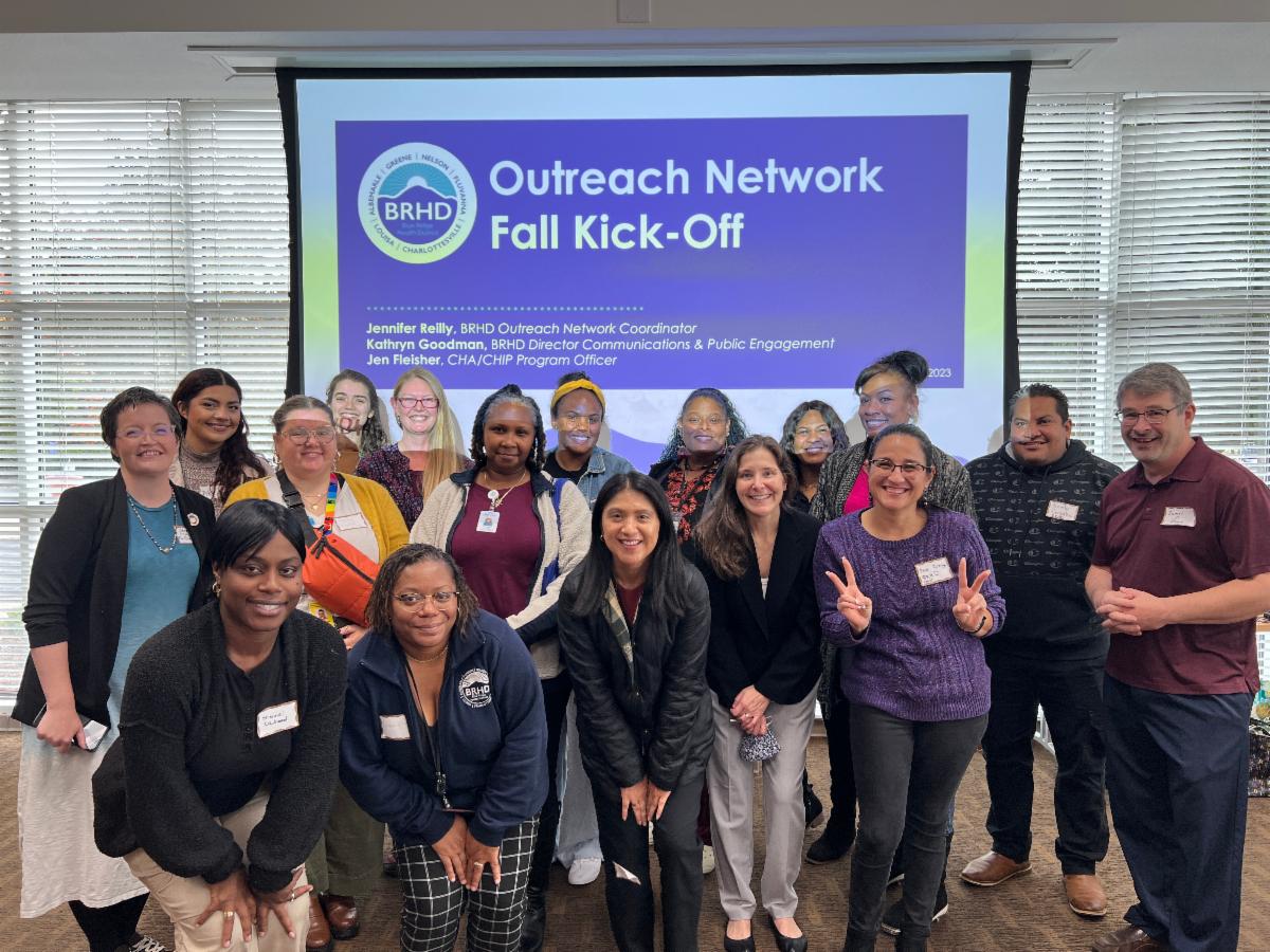 - 16 people wearing name tags and smiling pose in front of a powerpoint projection that reads “Outreach Network Kick-Off”.