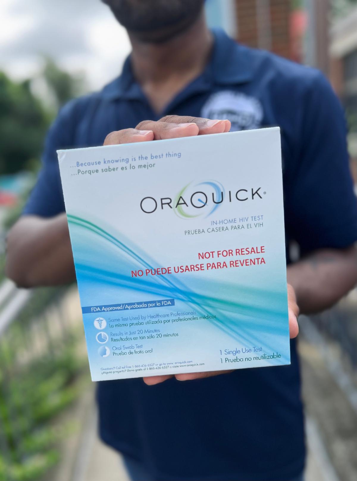 A black man in a navy blue BRHD polo slightly out of frame & focus holds up a white OraQuick brand rapid HIV Test Kit box up to the camera.