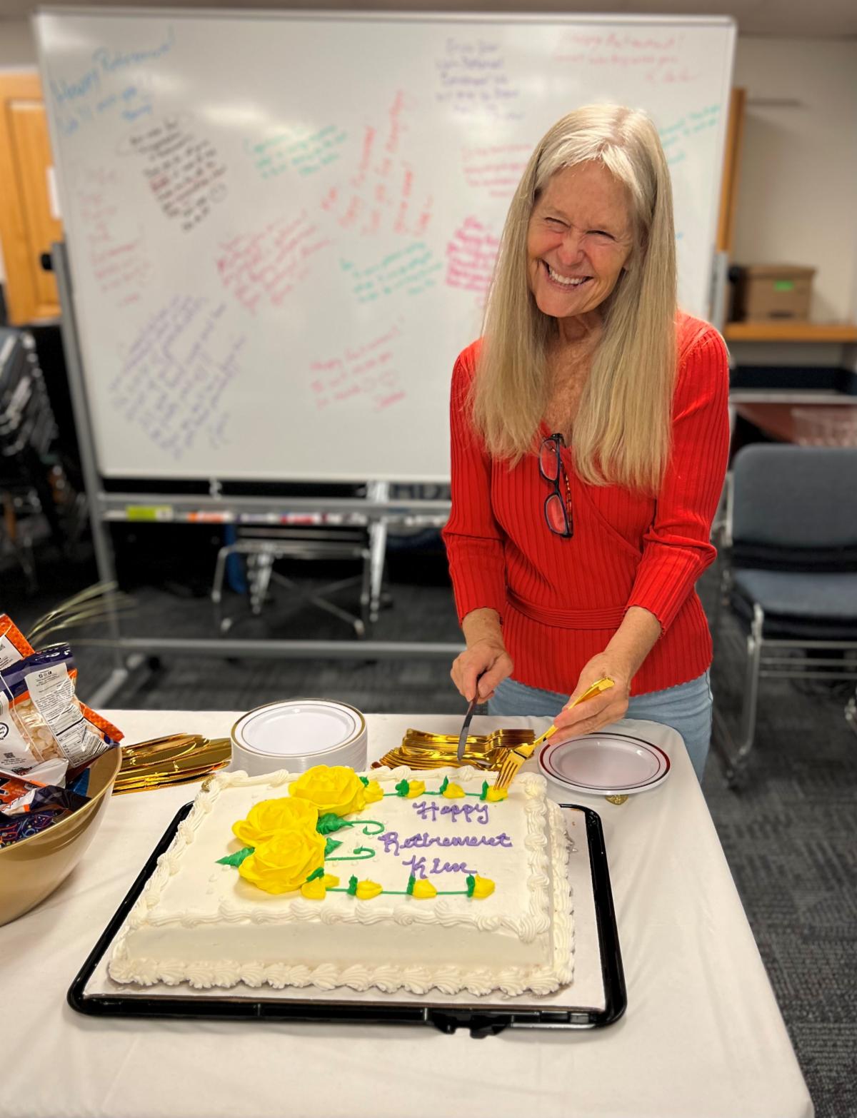 - Woman in a red sweater smiling at the camera while cutting a slice of sheet cake topped in yellow roses. The purple icing on the cake spells out “Happy Retirement Kim”. 
