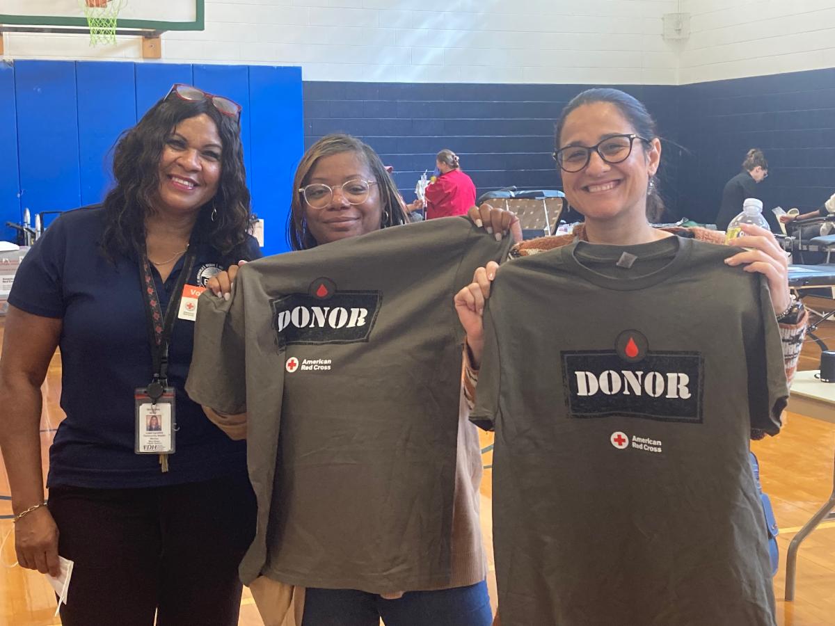 Inside a gymnasium three BRHD Community Health Workers stand side-by-side smiling at the camera. Two of the three CHWs are holding up gray and red blood donor t-shirts.