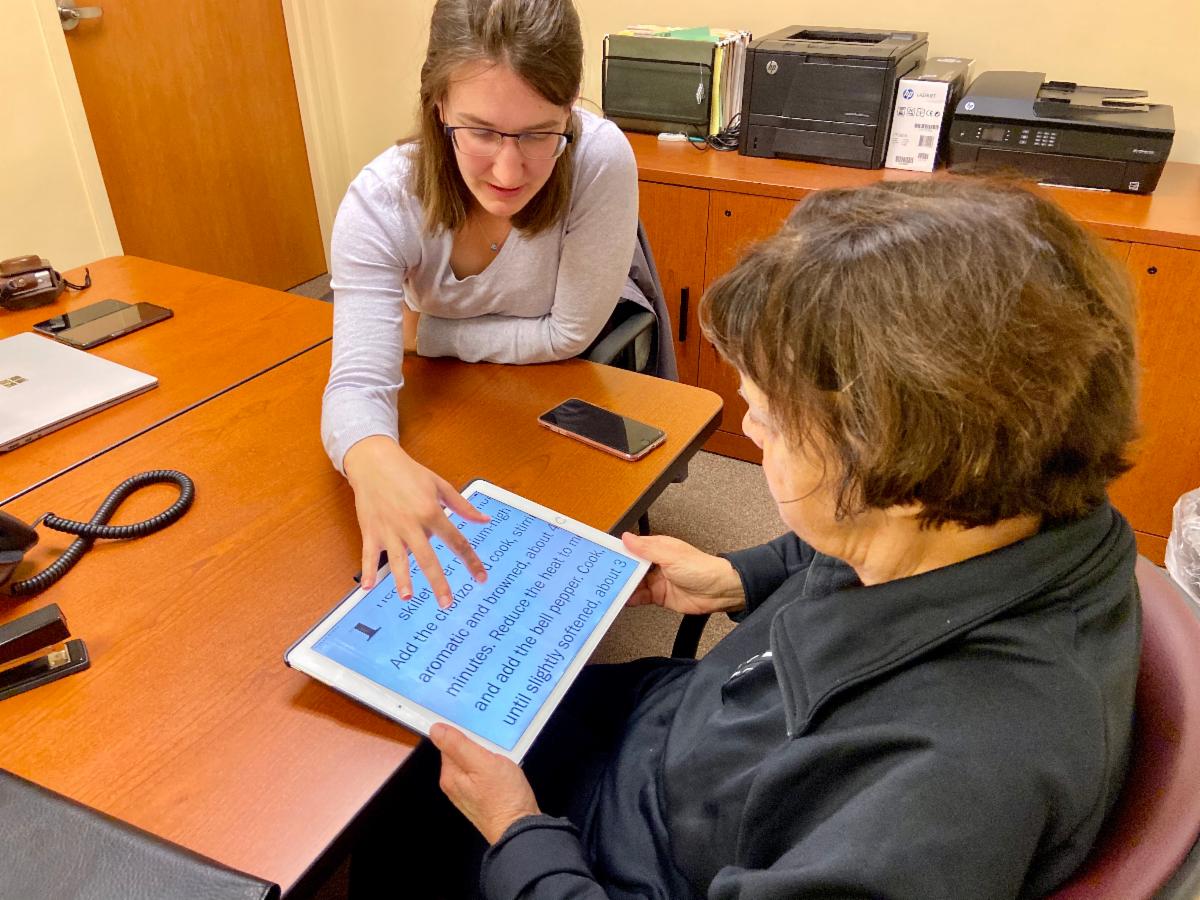 Two women sitting at a table. Woman on the left is demonstrating an iPad with enlarged text to a woman in the foreground
