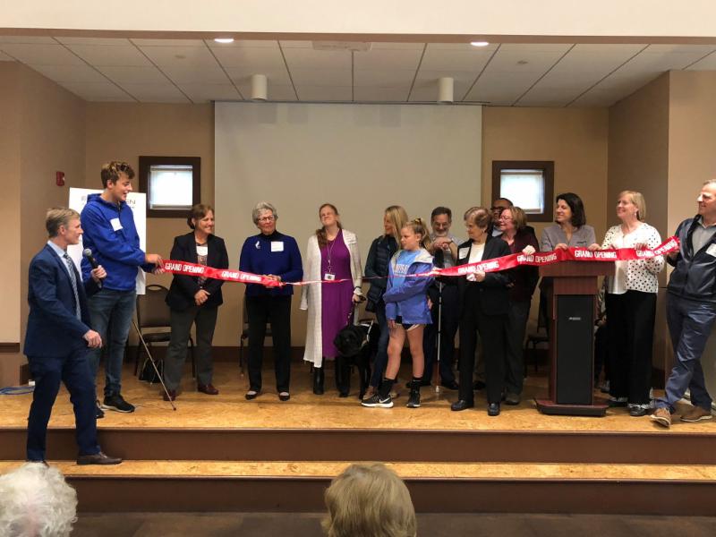 Elaine Ellenbogen standing on the stage surrounded my family cutting a big red ribbon. 