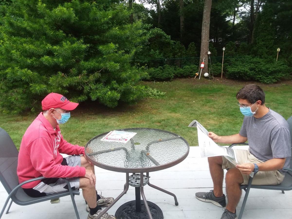 A gentleman wearing a pink sweatshirt and a mask sitting at a patio table facing a man in a mask reading a newspaper to him