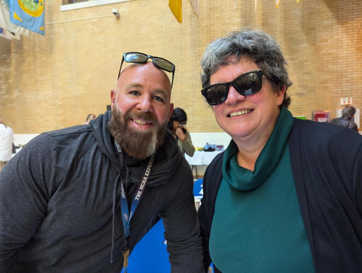 A man with a beard smiles next to a women with dark glasses.