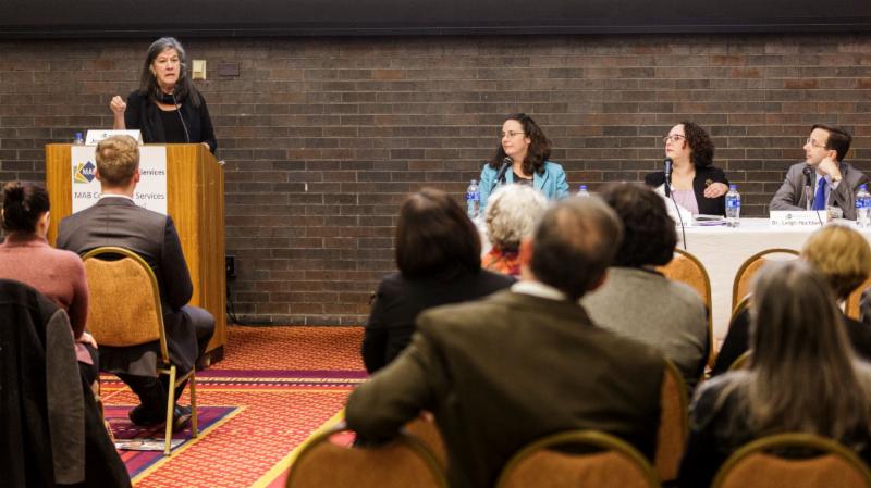 Left to right, board of director,Jean McGuire, standing at a podium with panelist, Kathryn Rucker, Amy Bernstein, and Dr. Leigh Hochberg 