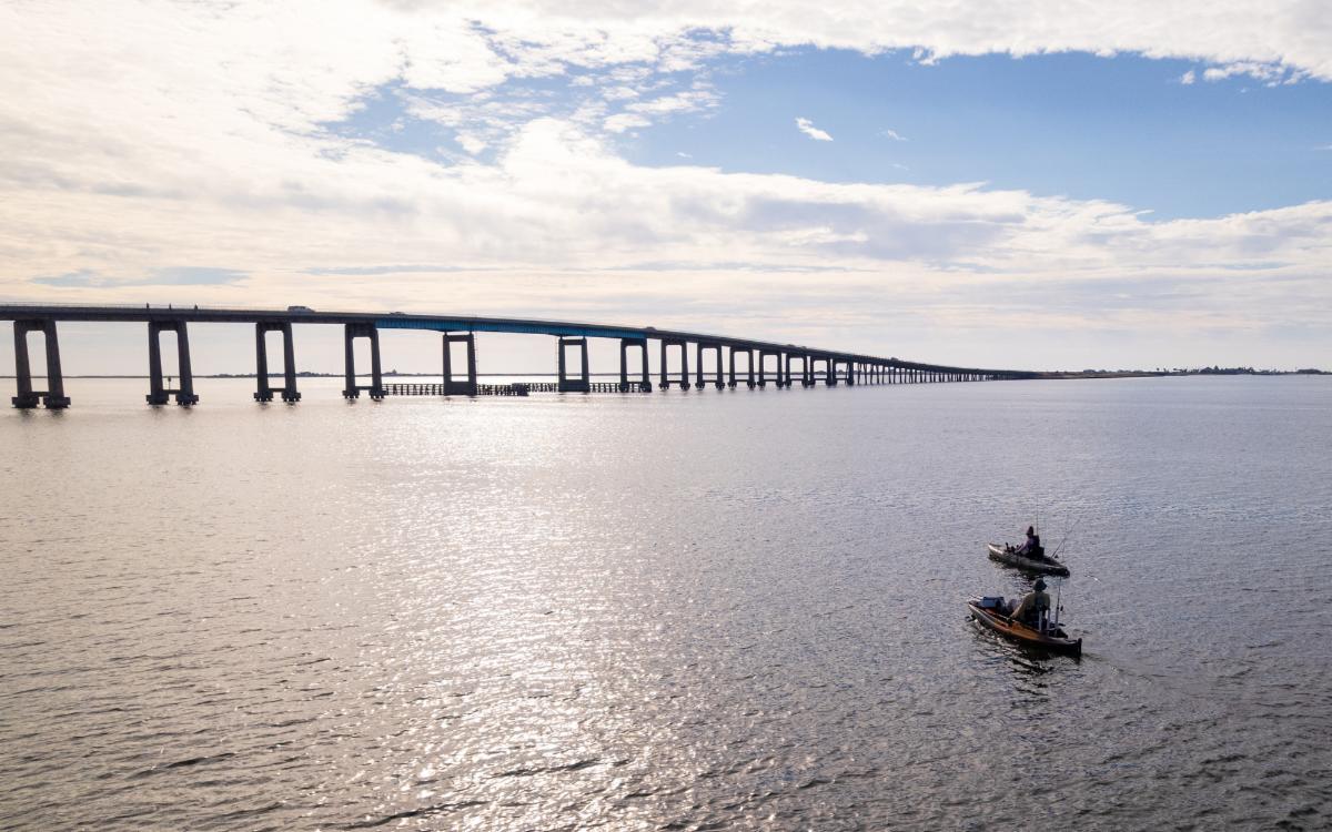 Kayakers on calm waters in the lower right hand corner. In the background is the Navarre Beach Bridge with a blue sky filled with white clouds behind it.