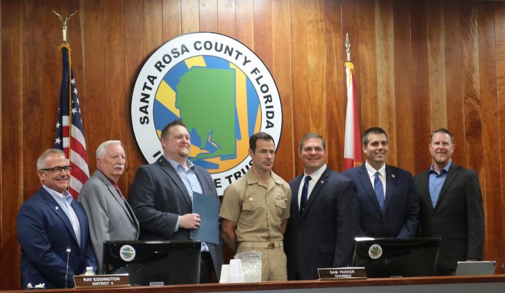 Board of County Commissioners standing in front of an American Flag and the County Seal. From left to right: Kerry Smith, Ray Eddington, Casey Powell, Captain Paul Flores, Sam Parker, James Calkins and Colten Wright