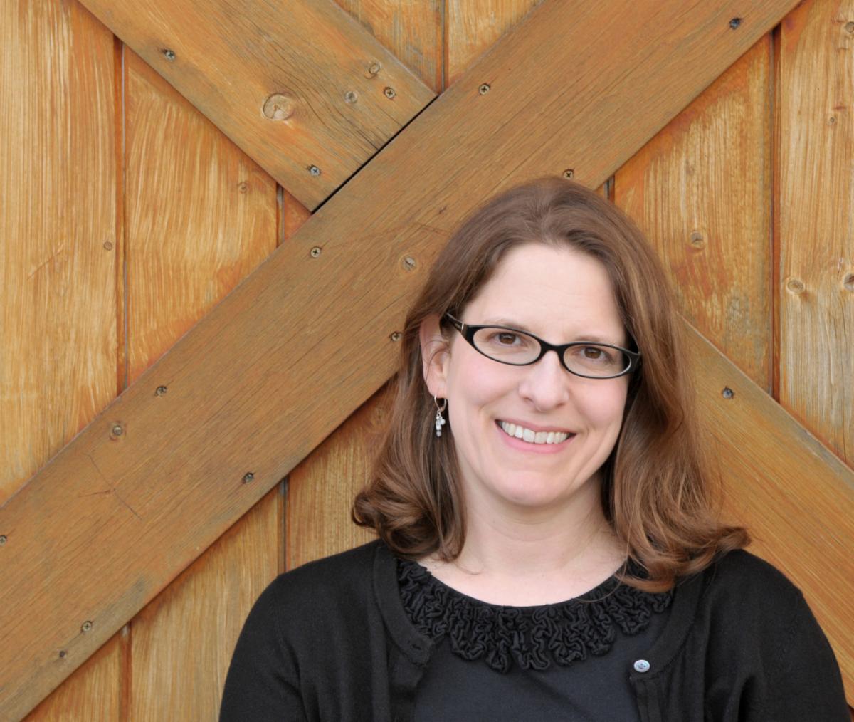 Photo of Kathy MacMillan in a black shirt and black glasses against a brown wooden door.