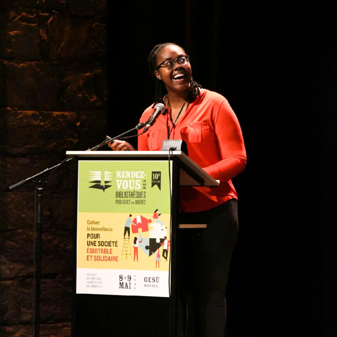 Photo of Kymberlee Powe presenting on a dark stage behind a podium as she smiles and looks up towards the presentation.