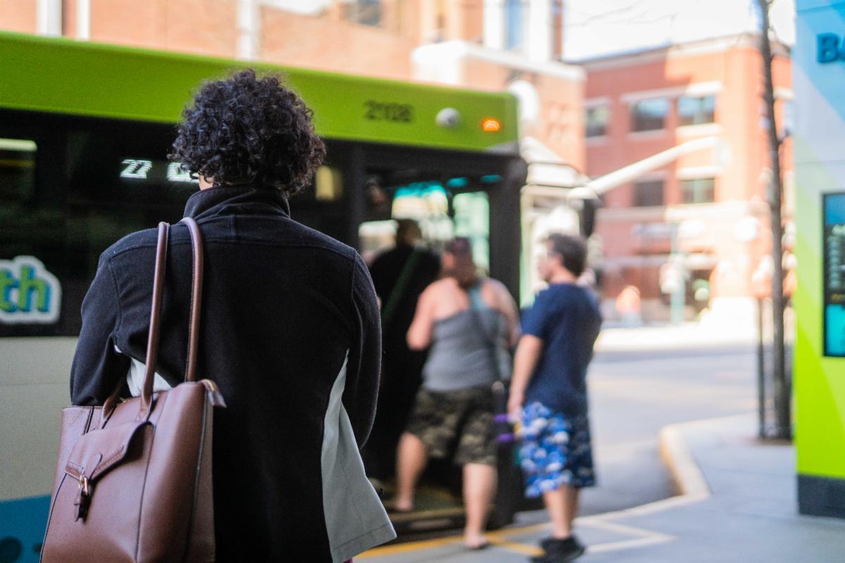 A photo of a woman waiting at a bus stop while others board a bus in the background