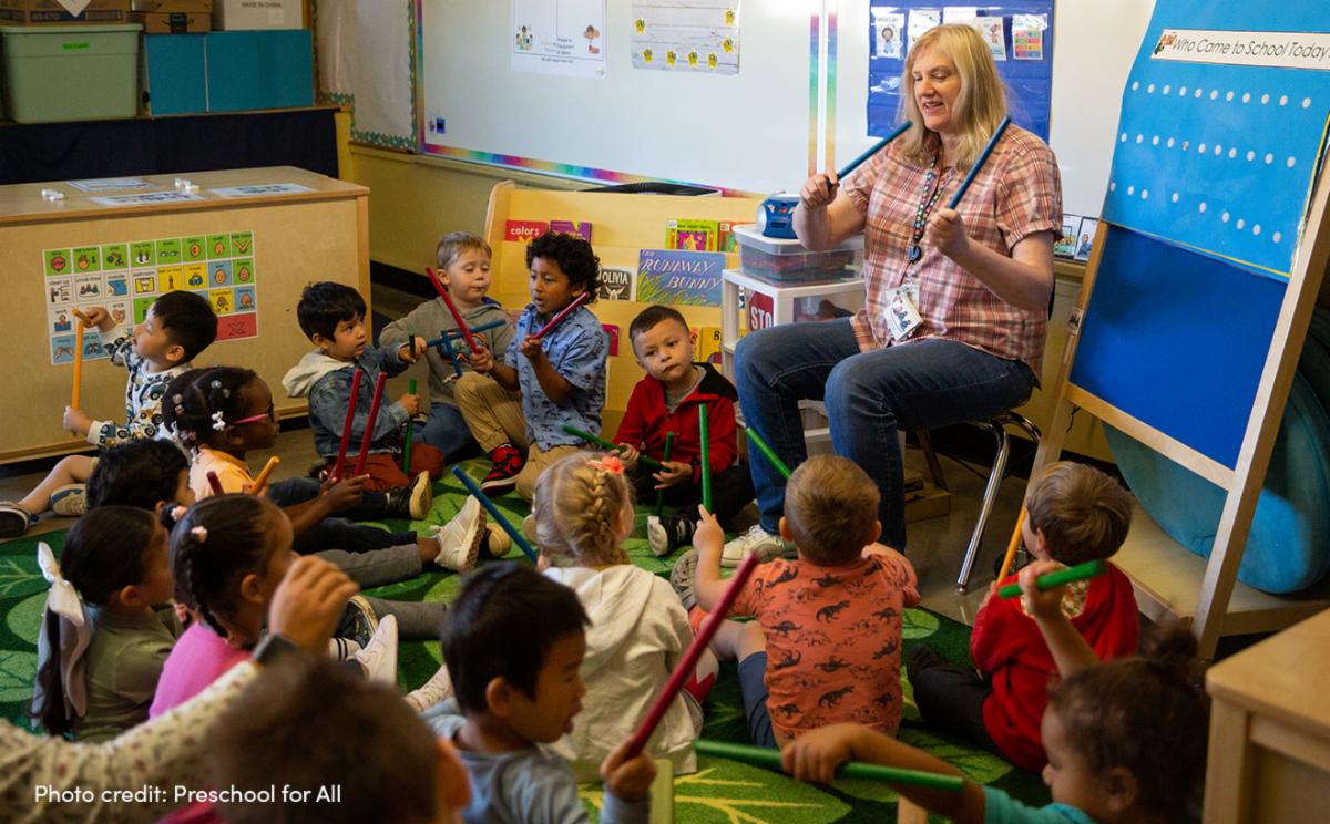 Preschool teacher leading kids in playing percussion instruments.