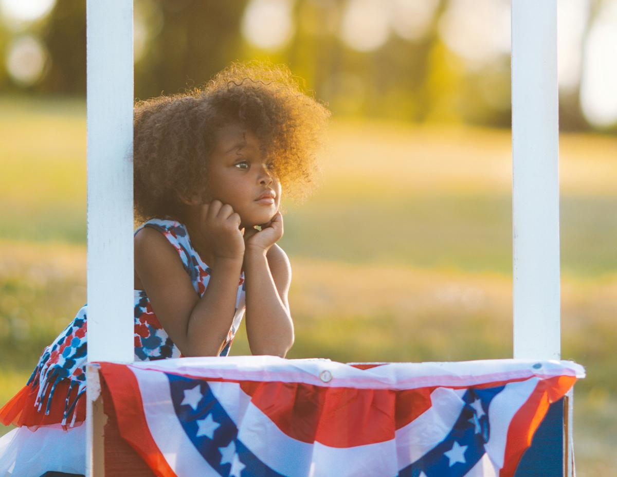 Child sitting behind a tractor steering wheel afront and American flag.