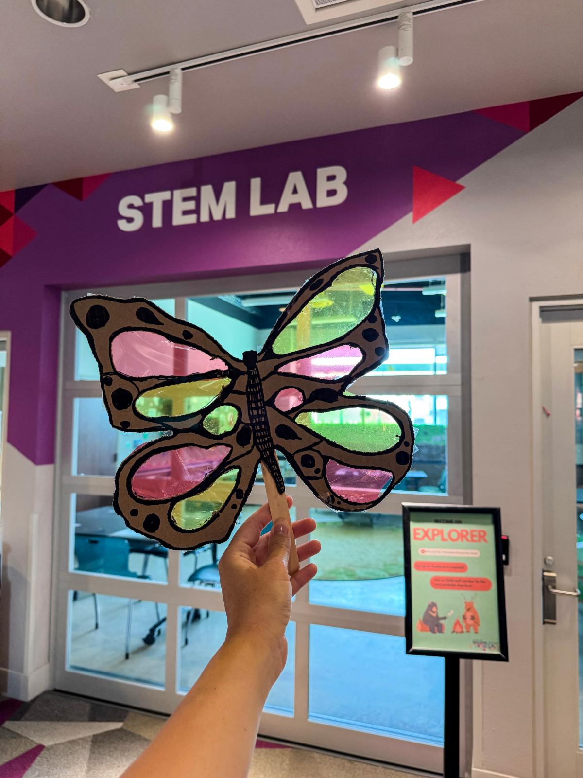 A person holds up a handmade butterfly craft made from cardboard and colorful translucent materials in front of the STEM Lab entrance at Randall Lewis Second Story and Beyond®. The butterfly features black outlines and decorative spots, demonstrating a creative project that can be made during the July cardboard makerspace activities.