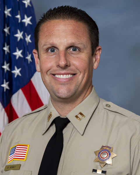 Head-and-shoulders portrait of a uniformed San Bernardino County Sheriff's captain Brad Bonnetsmiling at the camera. The captain is wearing a tan sheriff's uniform with captain insignia, a black tie, a department badge, and an American flag shoulder patch. An American flag is displayed in the background against a neutral gray backdrop.