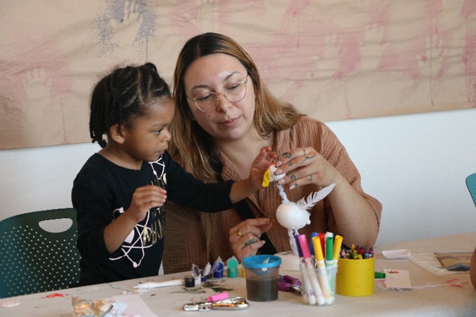 A photograph of a woman and a young child putting together a bird. On the table, there are multiple containers of paint of different colors, markers, and crayons.