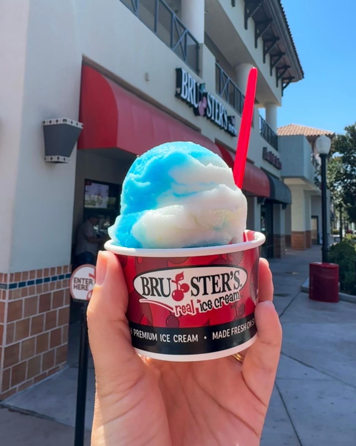 A person holds a cup of blue and white ice cream with a red spoon outside a Bruster's Real Ice Cream shop on a sunny day. The storefront and outdoor walkway are visible in the background.