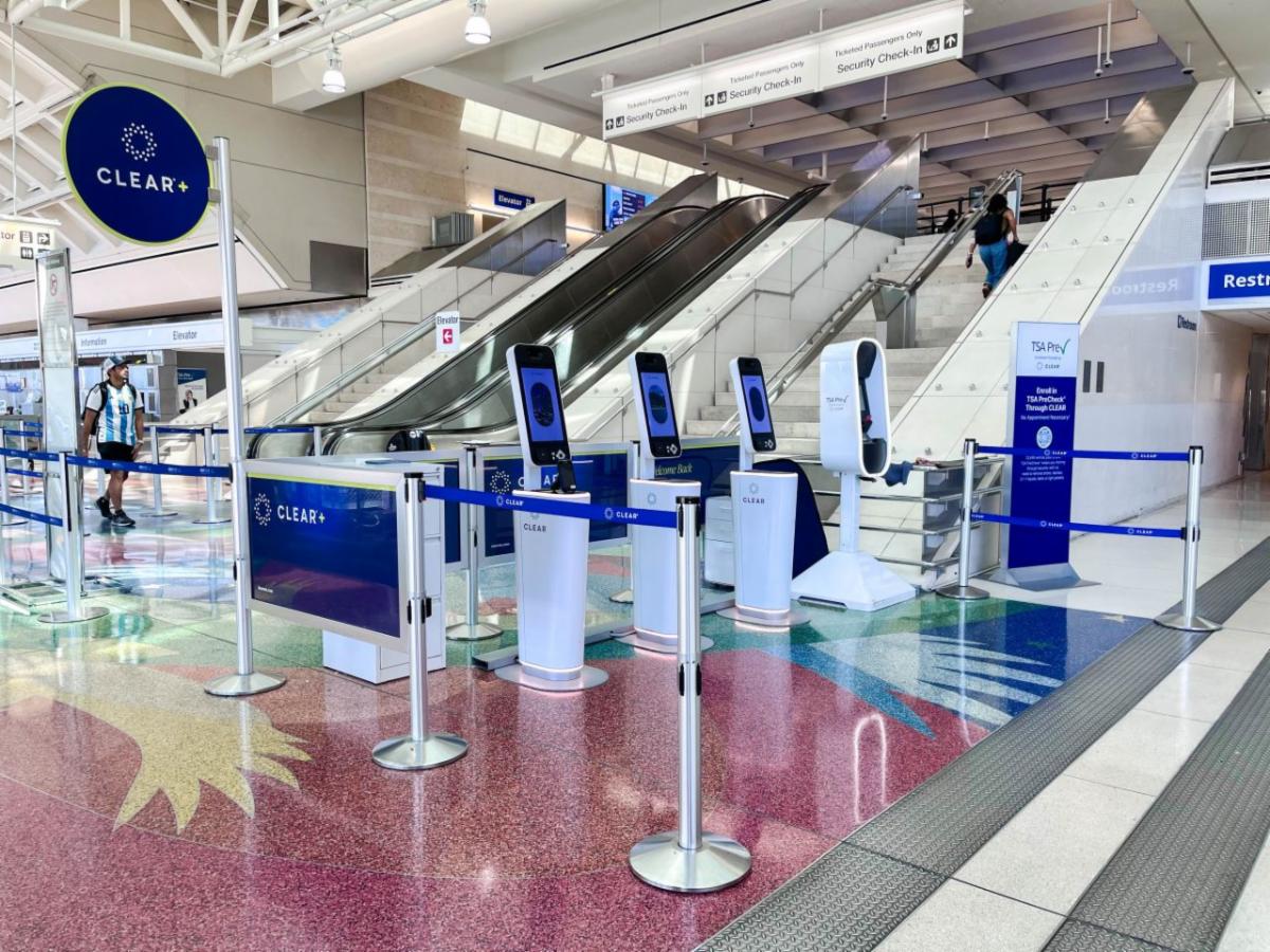 CLEAR+ identity verification kiosks and TSA PreCheck enrollment area inside an airport terminal, with security checkpoint escalators in the background.