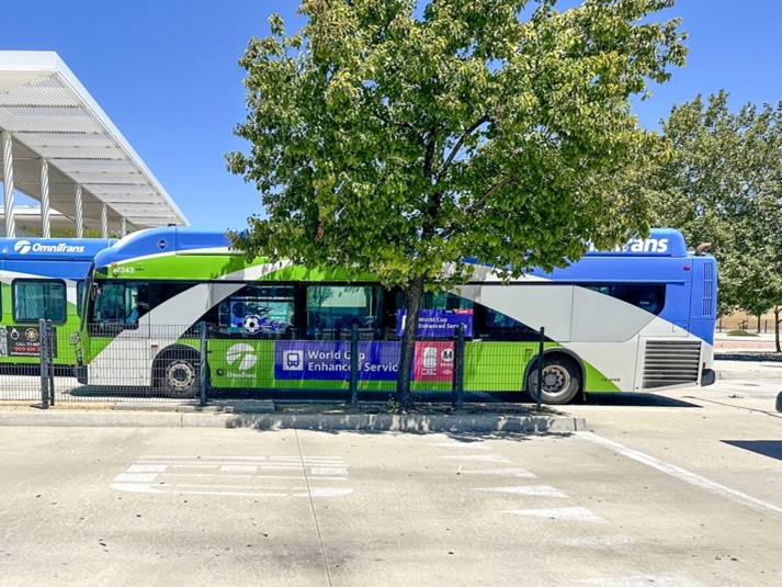An Omnitrans bus parked at a transit station behind a large tree, featuring promotional signage for World Cup enhanced service.