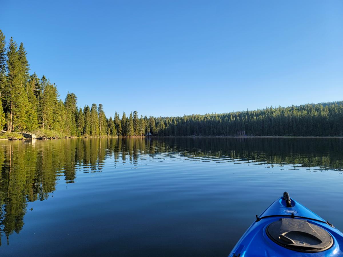 The font of a blue kayak looks out onto a calm glassy lake surrounded by pine trees