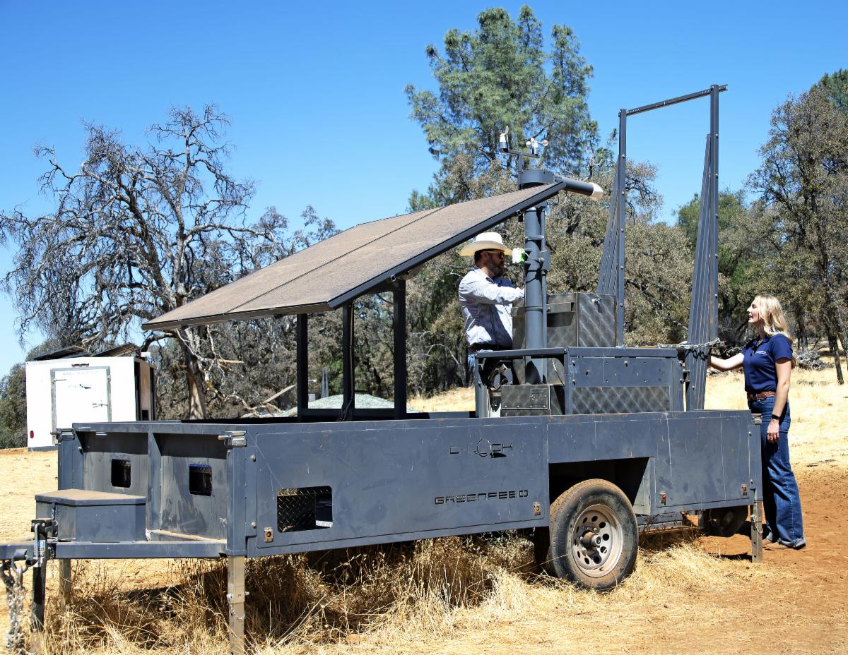 A woman stands next to a Green Feeder trailer, talking with a man wearing a cowboy hat who is in the trailer