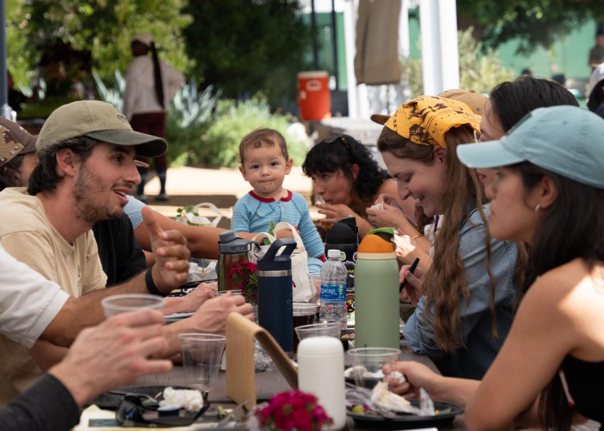Participants in an agritourism workshop sit and converse at a table while a young child looks directly at the camera