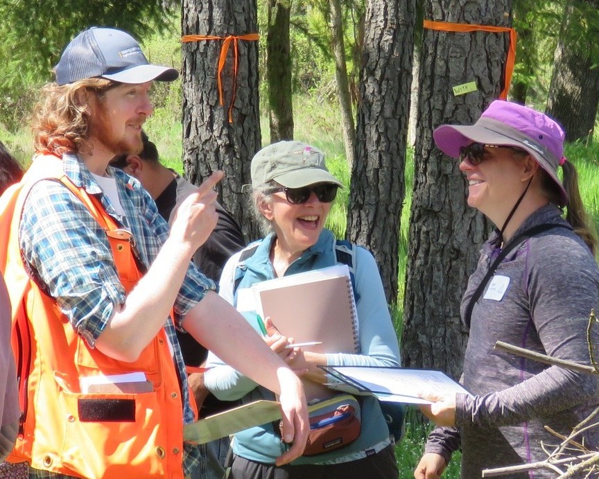 Instructor in blue UC ANR cap and orange vest gestures as he talks with two women wearing sunglasses amid a forest grove