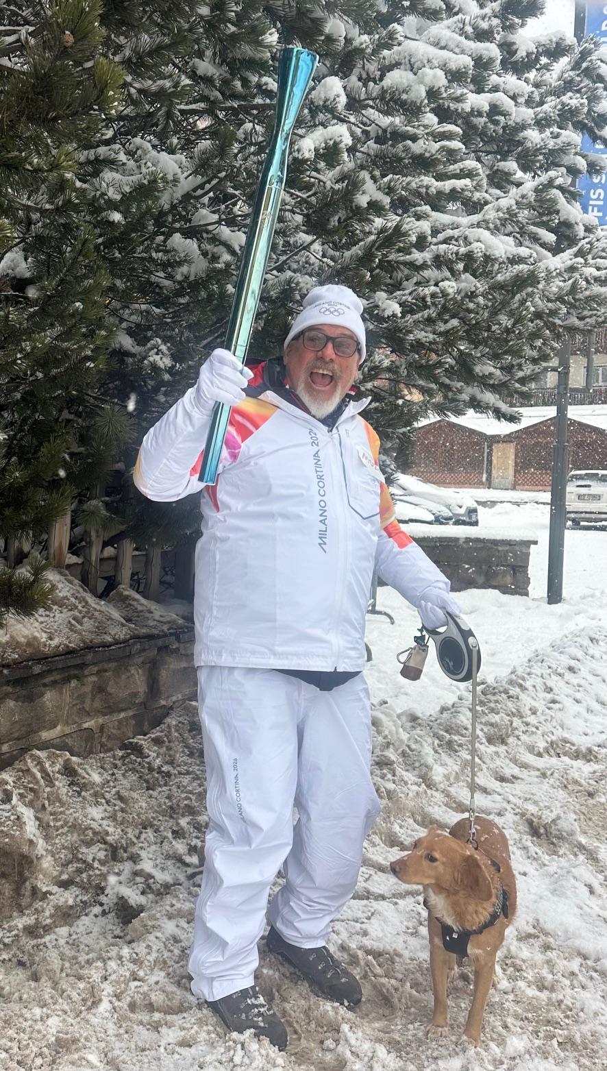 Man in a white parka and hat holds Olympic torch amid a snowy landscape, with a dog at his feet