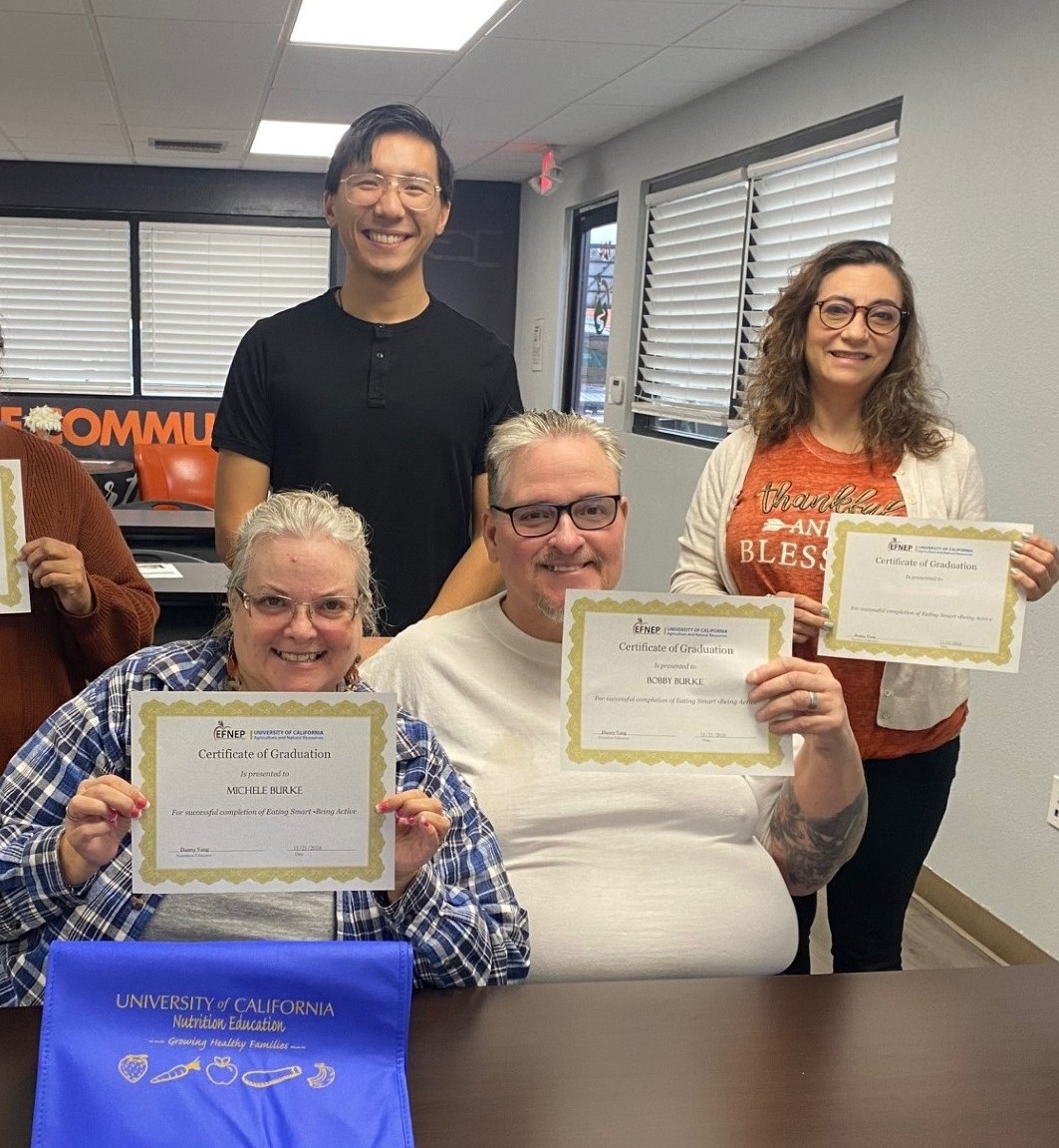 Three people hold up certificates of completion for a nutrition course as their instructor stands behind them in a classroom setting