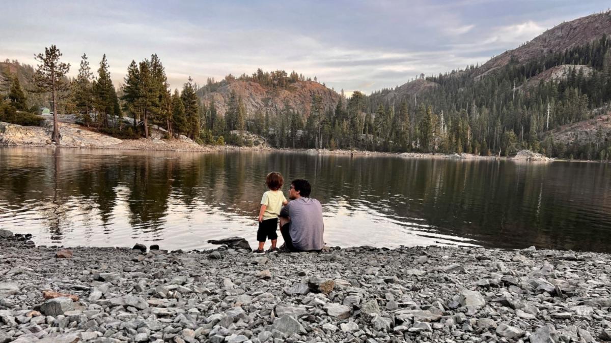 A man sits and a child stands next to him on the rocky shore of a lake, admiring the view of a mountain and its reflection in the water