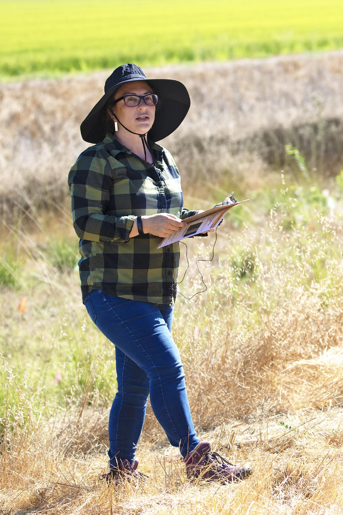 Woman in a wide brimmed hat holds a clipboard as she stands by rice fields giving a presentation