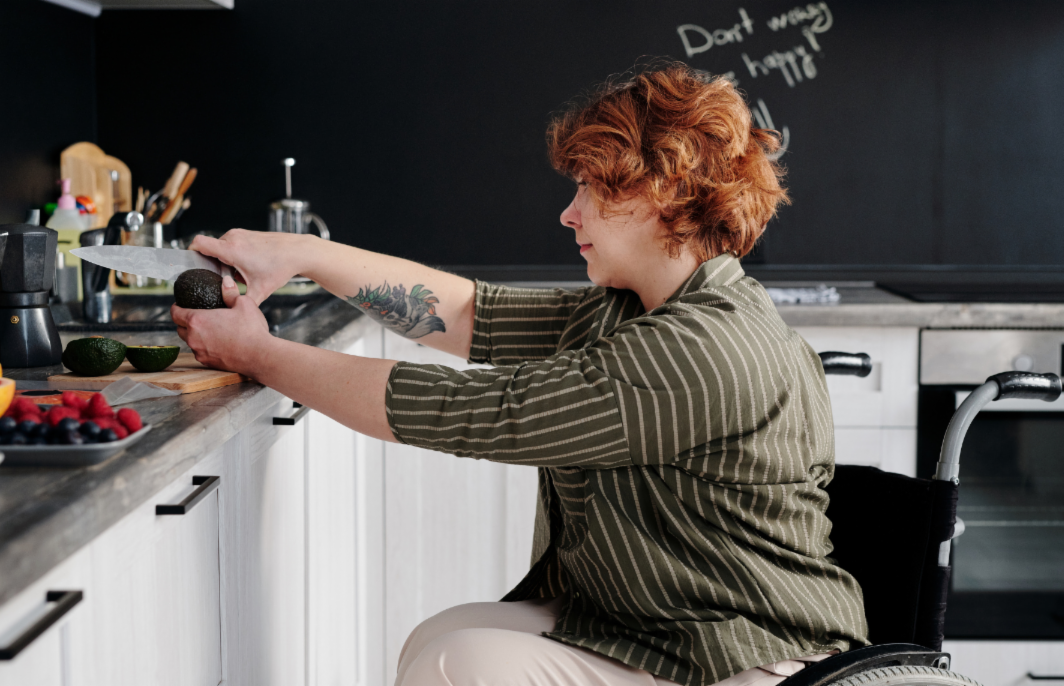 image of a female with short reddish brown hair, sitting in a wheelchair and leaning up to the kitchen counter with a knife in hand, cutting fruit to prepare a meal