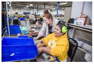 Female with low vision wearing a bright yellow sweatshirt sitting in a chair in a production facility assembling products for packaging 