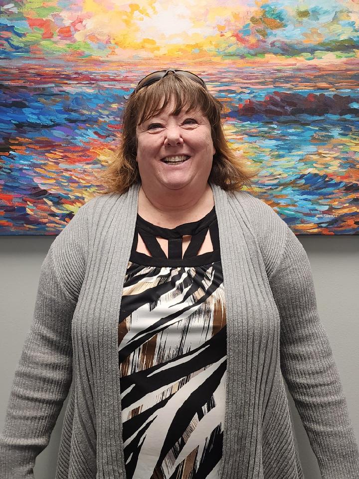 Female with shoulder length reddish brown hair wearing a gray cardigan standing in front of a colorful painting smiling at the camera