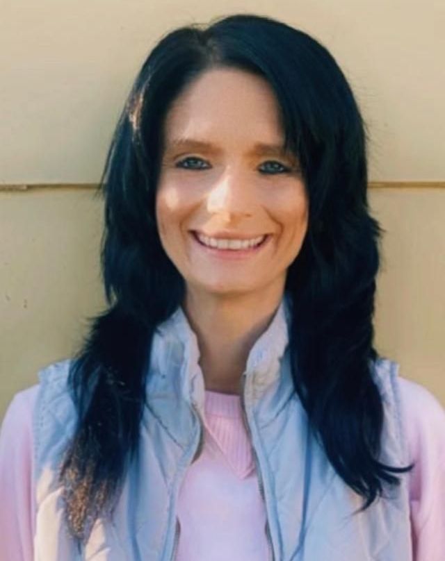 closeup photo of a light skinned female with long black hair wearing a pink shirt and light blue vest smiling at the camera