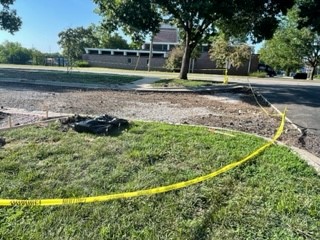 image of a portion of a parking lot that is under construction. In the forefront is a patch of grass and in the background is a building. A section of yellow caution tape is pictured partially surrounding the construction area.
