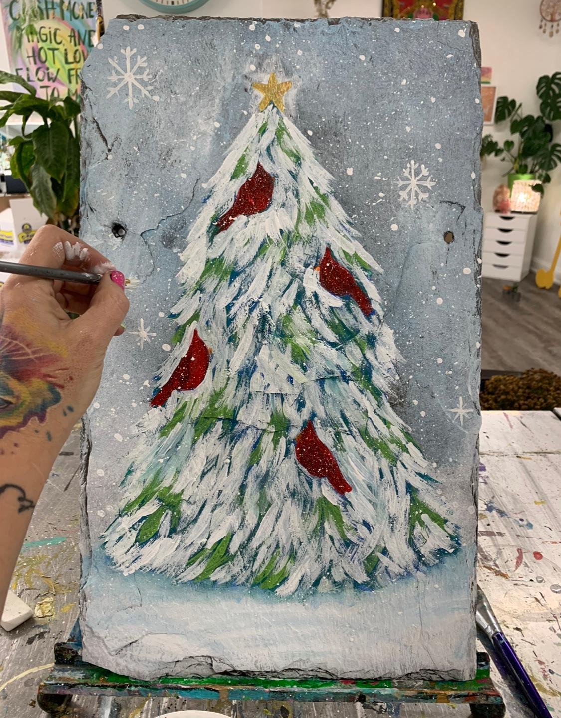 A hand painting a snow covered evergreen tree with 4 cardinal birds on it.
