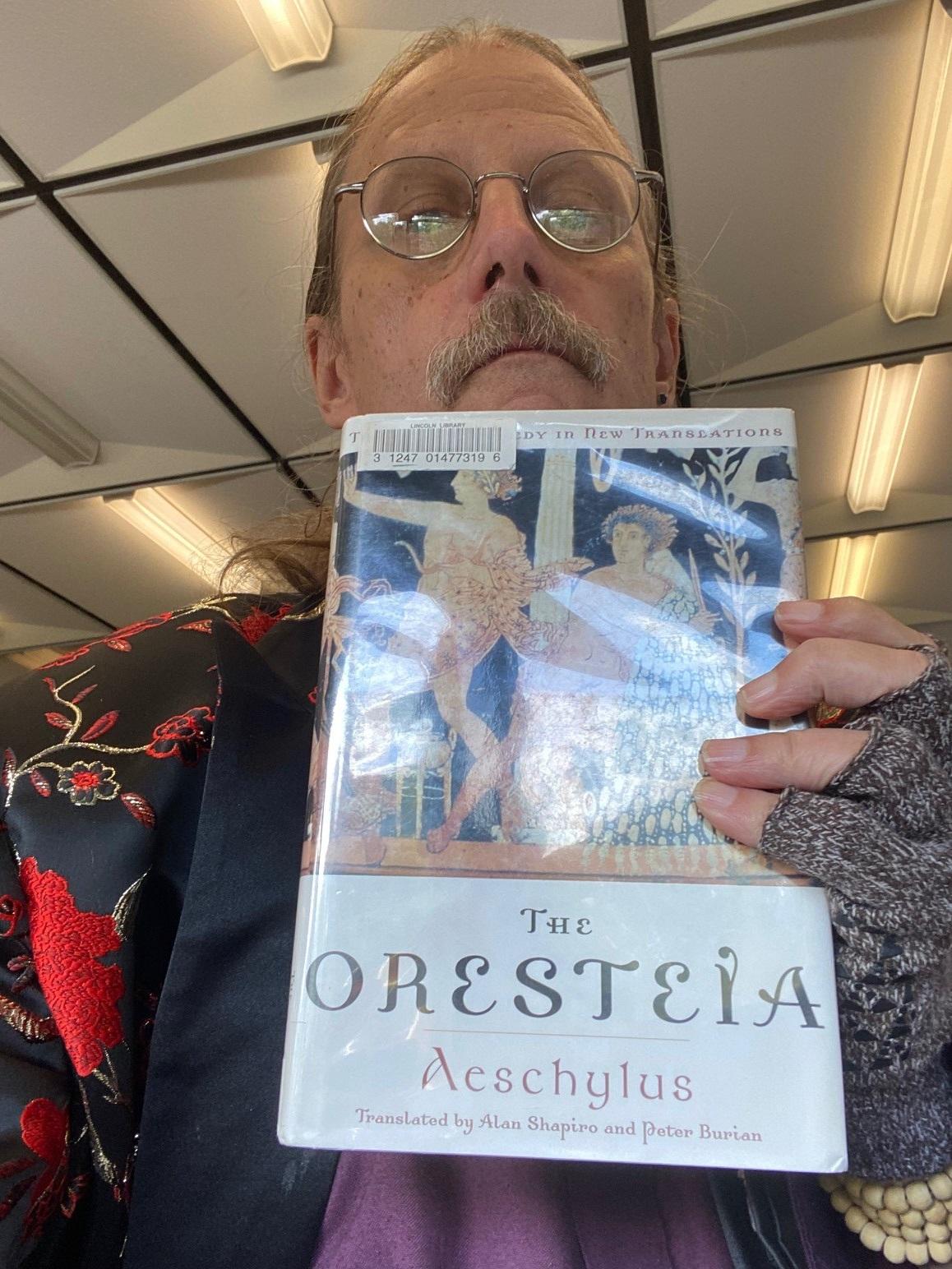 A man with glasses and a moustache holds a copy of The Oresteia