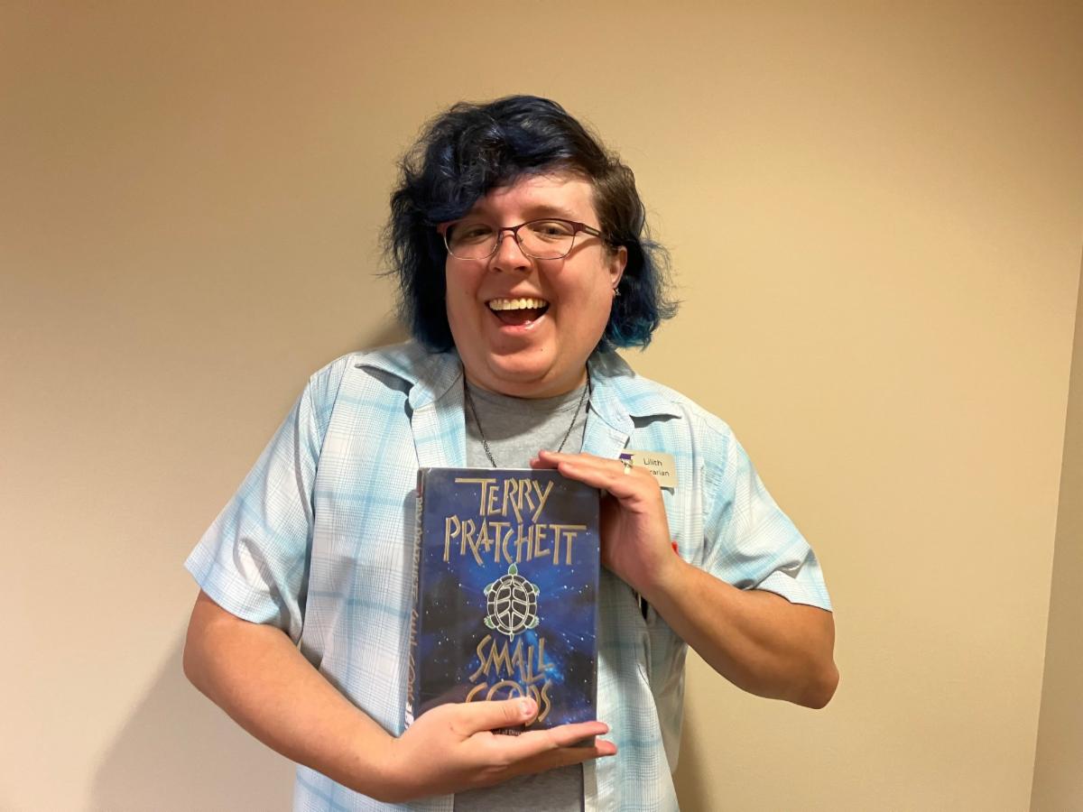 A smiling woman with blue hair holds Small Gods by Terry Pratchett