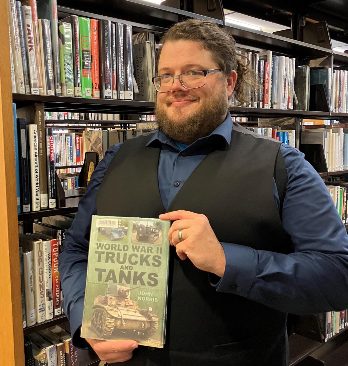A man stands in front of bookshelves holding a copy of the book "World War II Trucks and Tanks" 