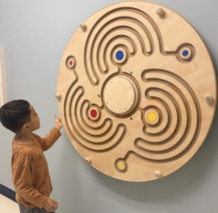 young boy with dark hair looking at a tactile wooden wall sculpture.