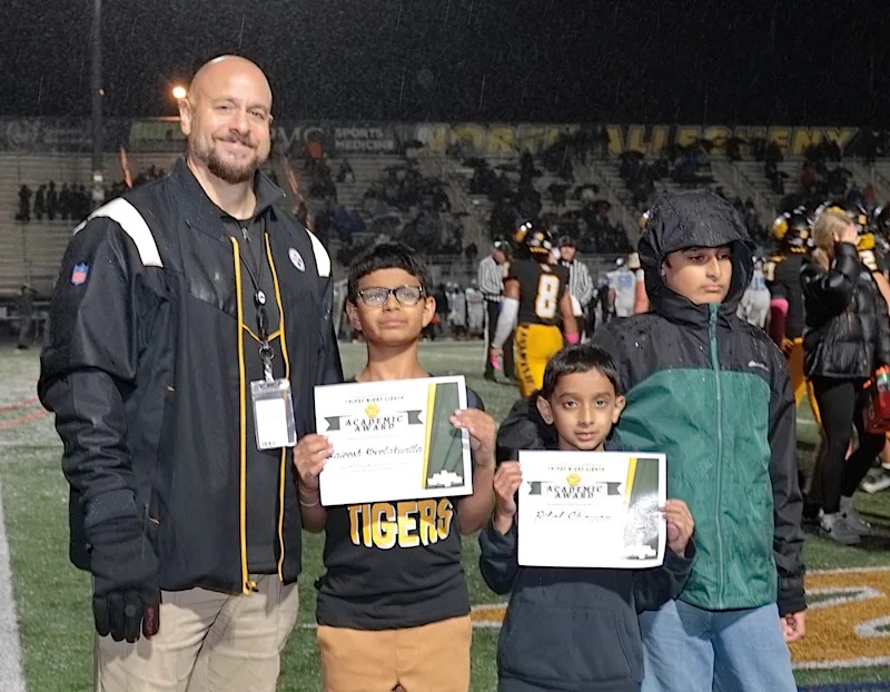 North Allegheny Superintendent Mr. David Deramo standing on a football field alongside two elementary students displaying their Friday Night Lights Academic Award certificates.