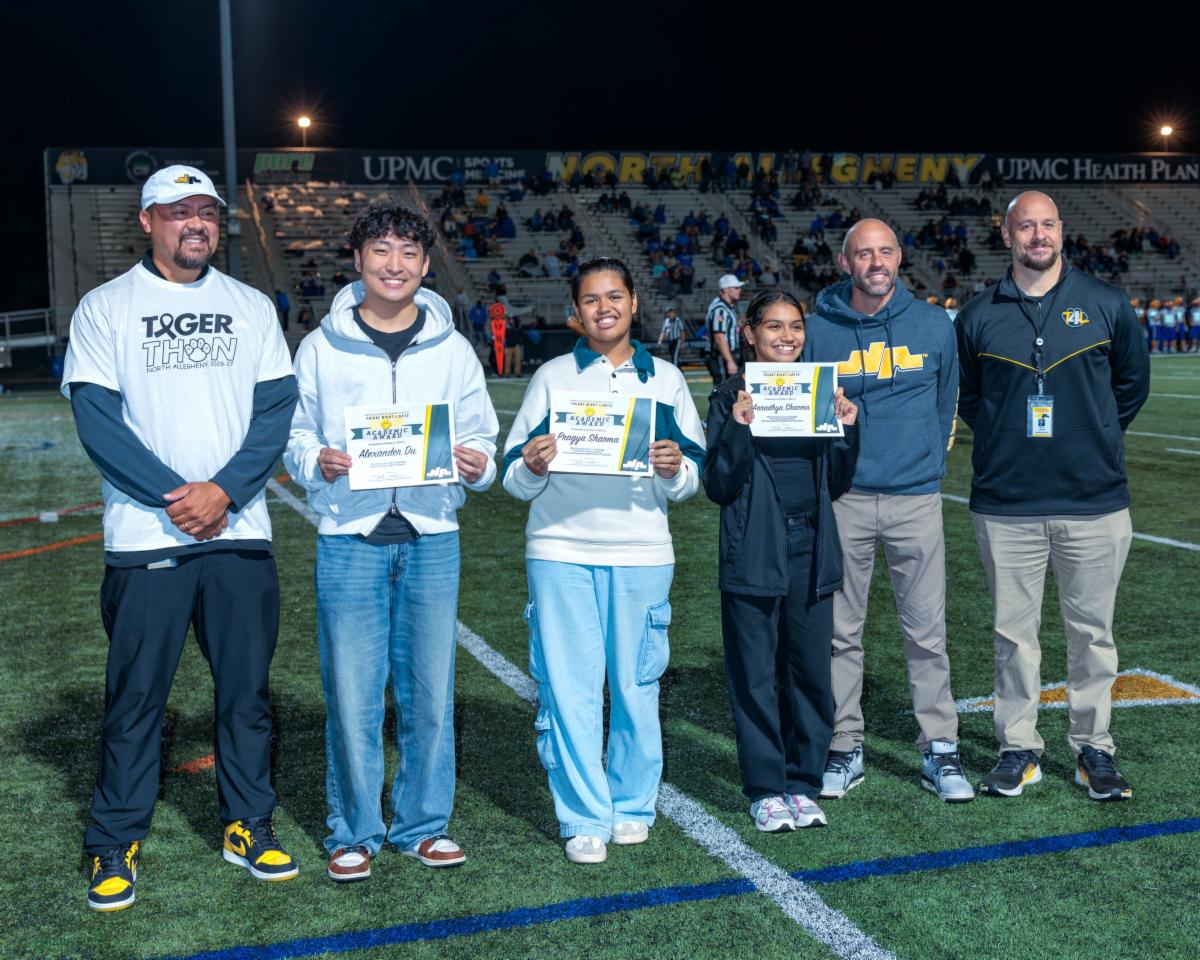 Six people stand on a football field track at night posing for a photo. In the center, three North Allegheny students hold up official certificate papers displaying their names: Alexander Du, Pragya Sharma, and Aaradhya Sharma. They are flanked by three school administrators. The group is smiling on the green turf beneath stadium lights, with bleachers and North Allegheny signage visible in the background.