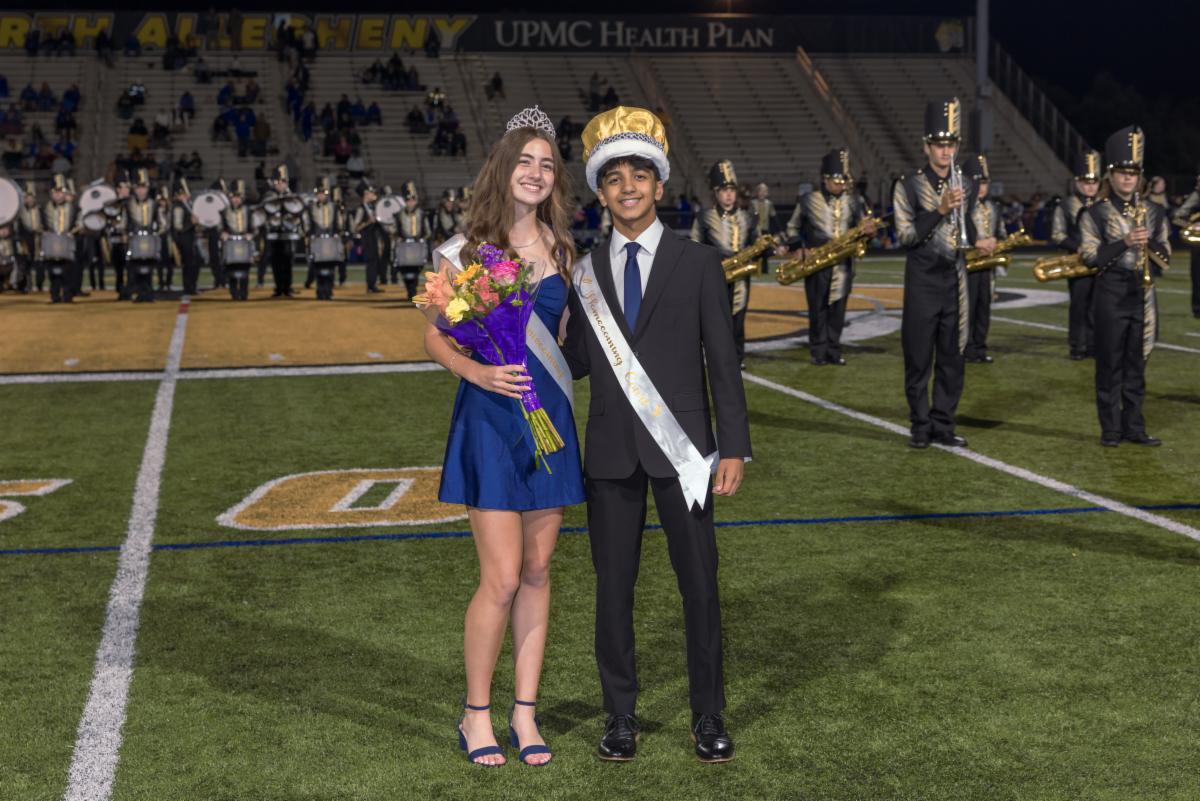 North Allegheny Homecoming king Arjun Puri and queen Addison Goetz stand smiling on the football field. Addison wears a tiara, blue dress, sash, and holds a bouquet of flowers, while Arjun wears a gold plush crown, suit, and sash, with the marching band lined up behind them.