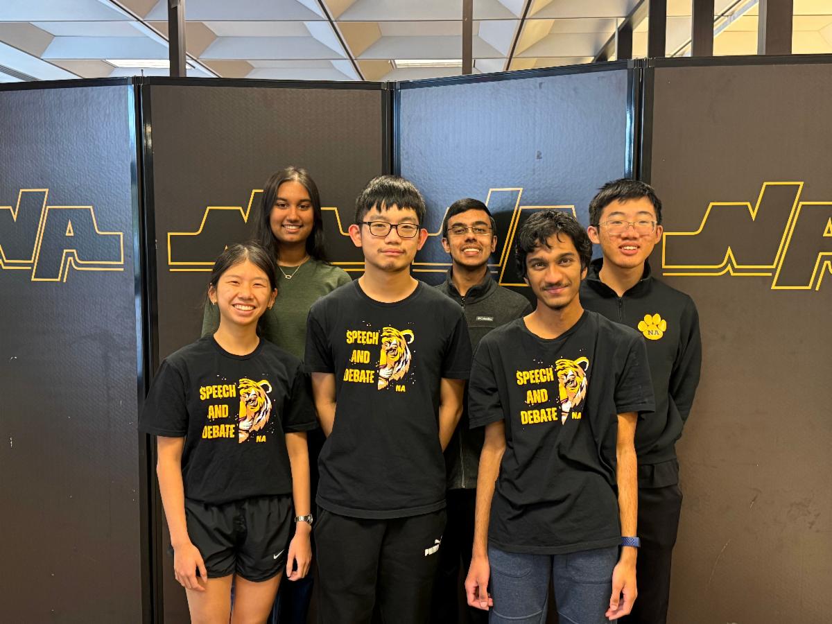 Six members of the North Allegheny Speech and Debate team standing together in front of a black backdrop featuring the gold "NA" school logo. Three students in the front row wear black "Speech and Debate" T-shirts featuring a tiger mascot. The group is smiling, posing for a commemorative photo following their qualification for the International Public Policy Forum Round of 32.