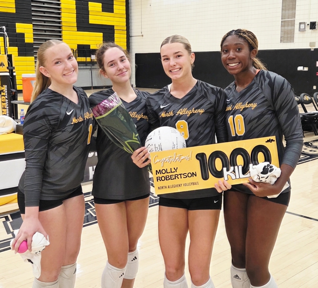 Four high school volleyball players in matching black patterned jerseys pose together on the court. The center player holds a white signed volleyball, a bouquet of wrapped flowers, and a large yellow and black sign celebrating her 1,000th career kill.
