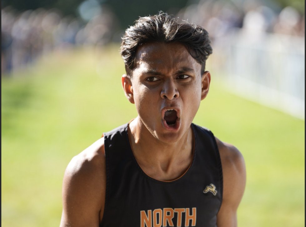 Close-up action shot of North Allegheny cross country runner Gatik Hirwani reacting with an intense, triumphant expression as he crosses the finish line. He is wearing a black North Allegheny singlet and is outdoors on a grassy course with a blurred background.