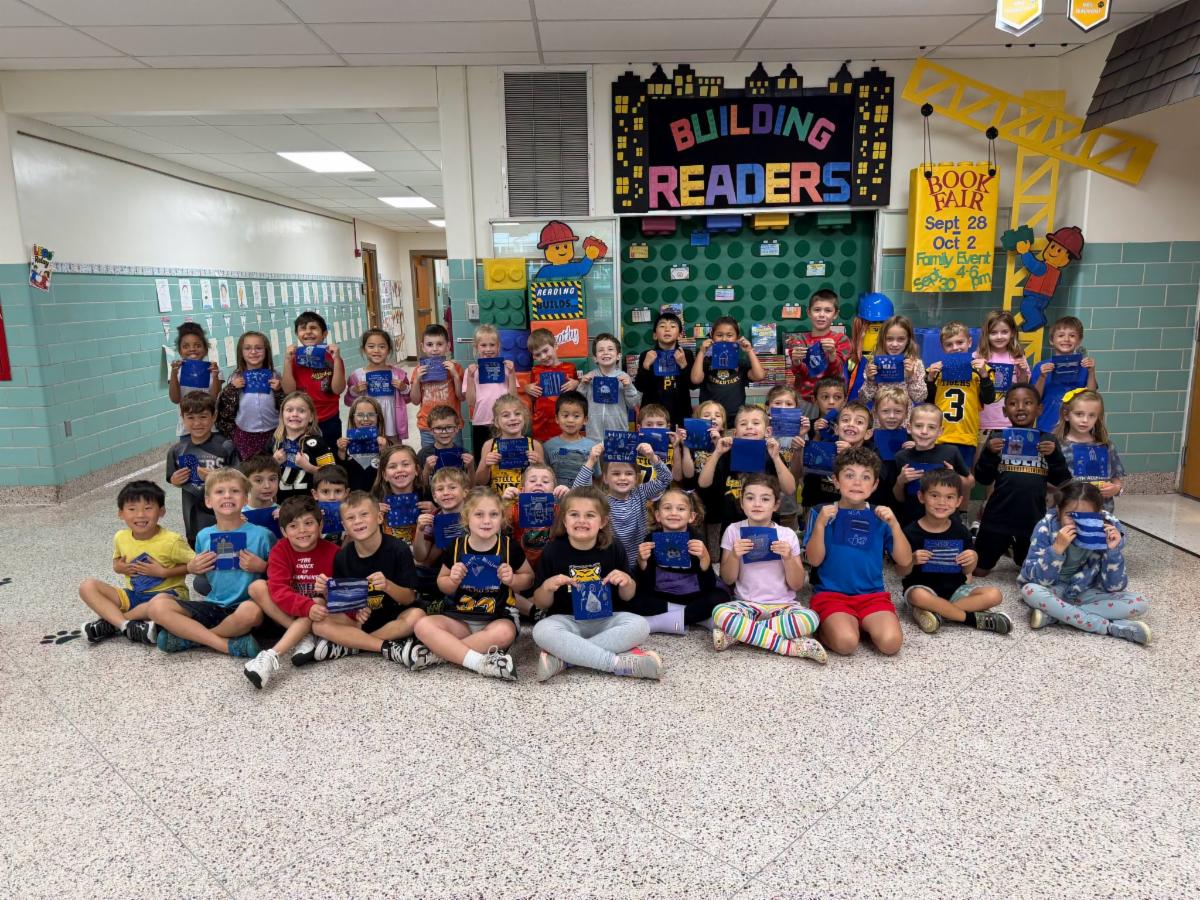 A large group of elementary school students poses together in a school hallway, seated on the floor and standing in rows. Each child holds up a small, decorated blue fabric tile in front of them. In the background, a colorful bulletin board reads "Building Readers" alongside construction-themed decorations and school announcements.