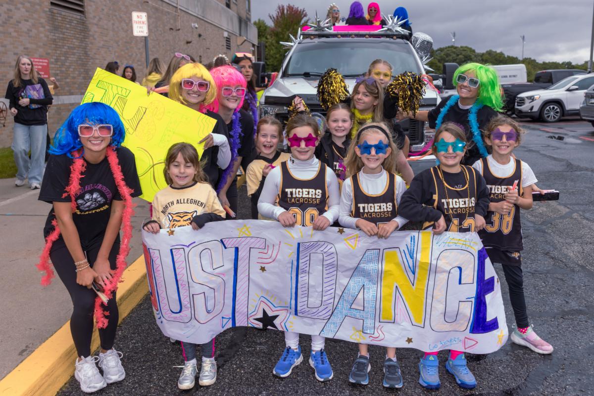 A group of young students and older cheerleaders/participants pose outdoors during the North Allegheny Homecoming Parade. The students in the front hold a hand-drawn sign that reads "JUST DANCE". Many in the group are wearing colorful neon wigs, star-shaped sunglasses, and North Allegheny Tigers Lacrosse jerseys, with a parade vehicle visible directly behind them.