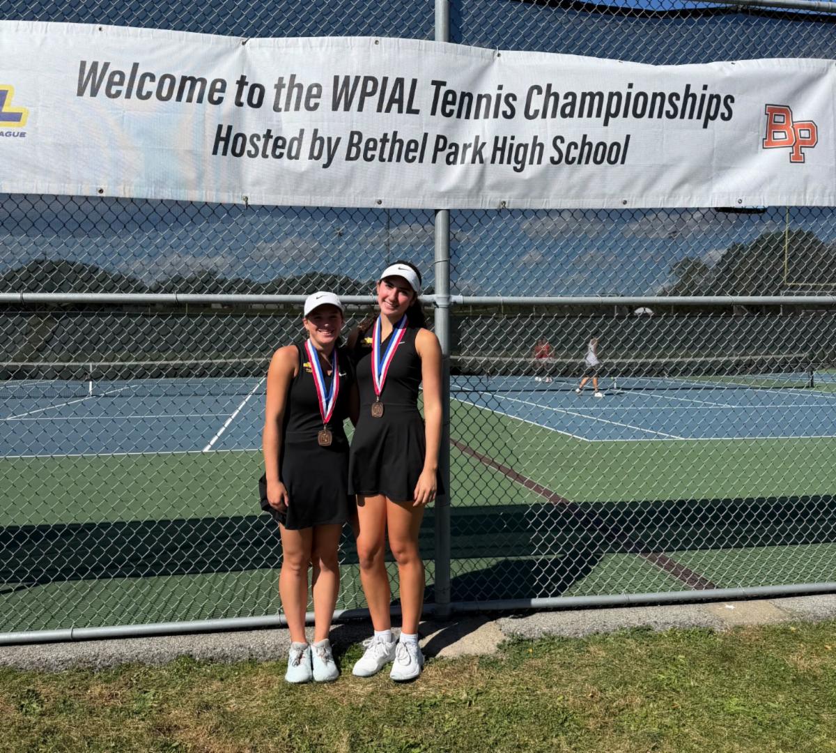 North Allegheny tennis players Mia Kaufman and Sammy Leeman smile together on a tennis court, wearing matching black uniforms, visors, and bronze medals. Behind them, a banner on the chain-link fence reads "Welcome to the WPIAL Tennis Championships Hosted by Bethel Park High School".